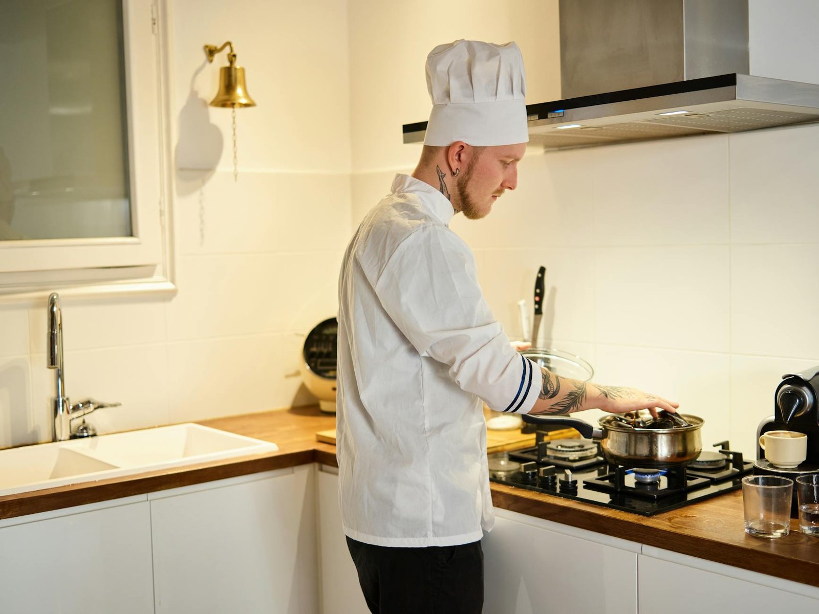 A tattooed chef wearing a hat cooks in a modern kitchen, focusing on a pot on the stove.