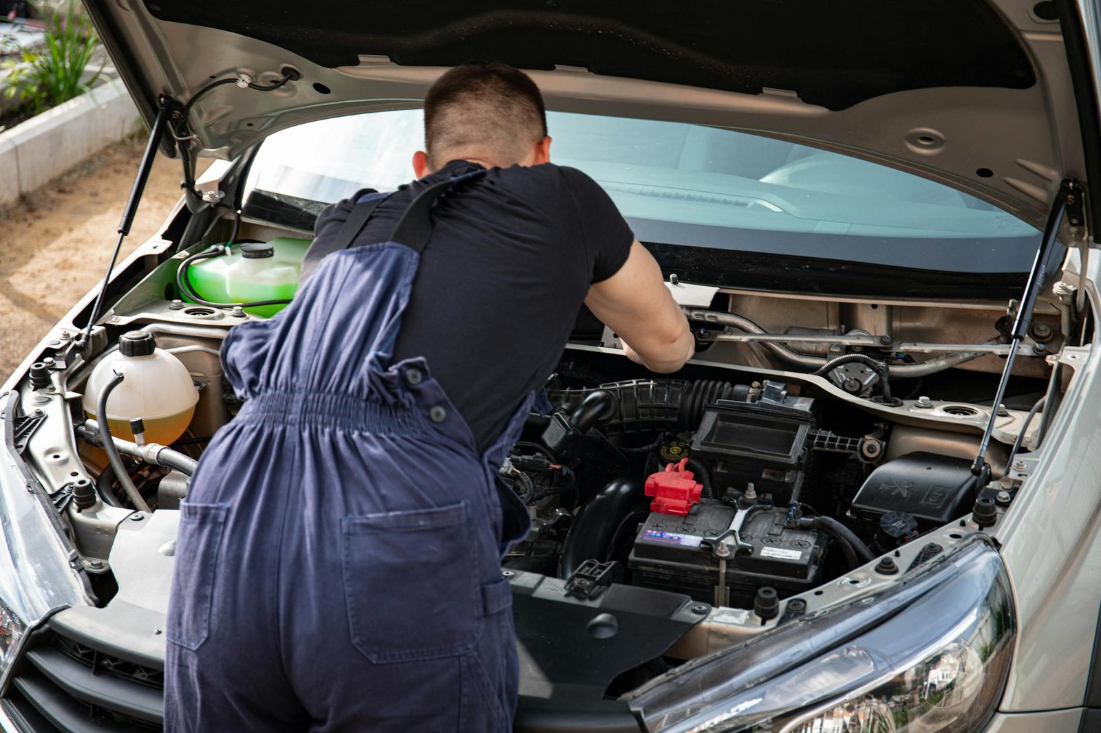Mechanic in blue coveralls performing car engine maintenance outdoors.