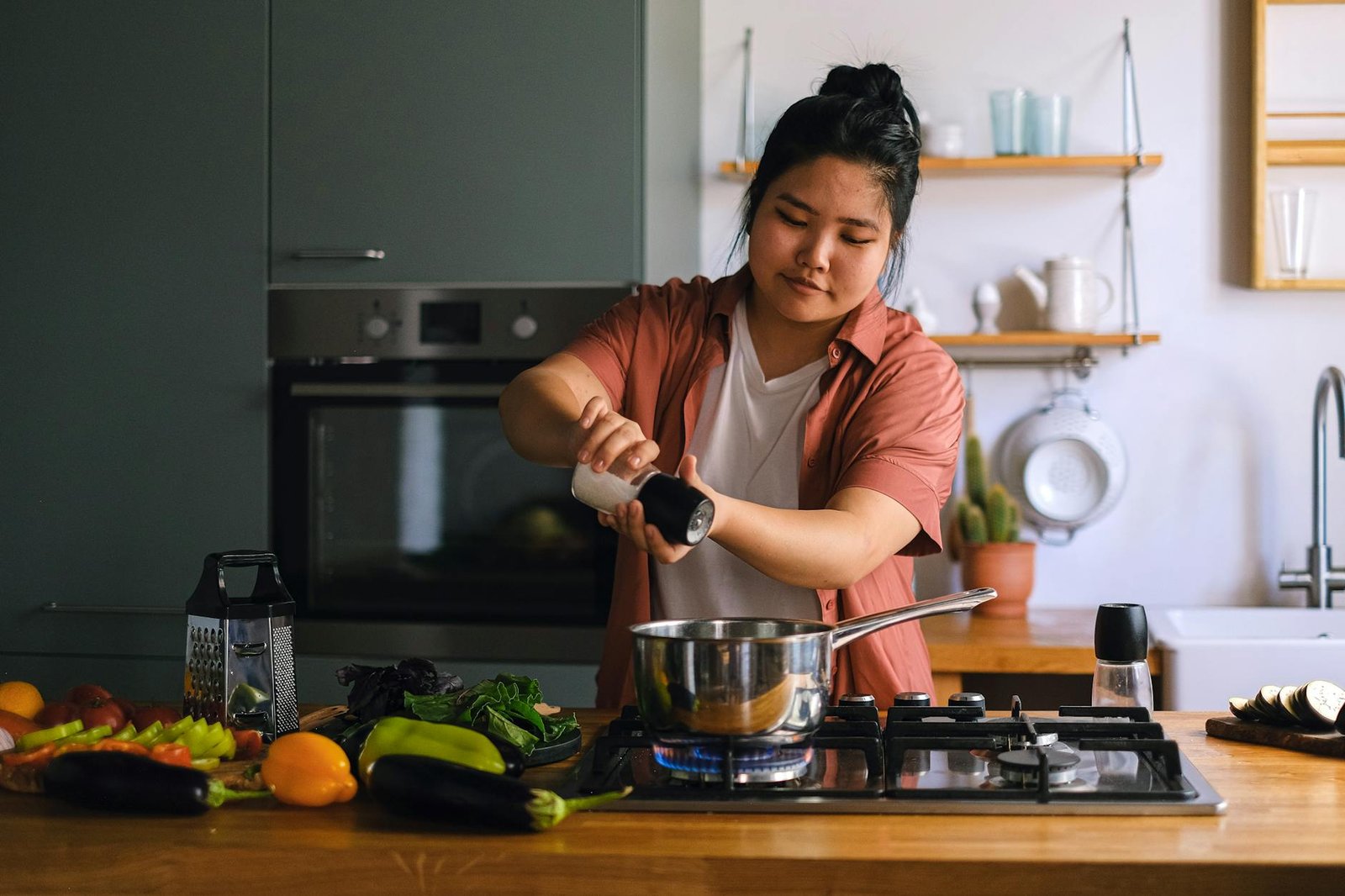 A home cook seasoning a dish on a stove surrounded by fresh vegetables in a modern kitchen.