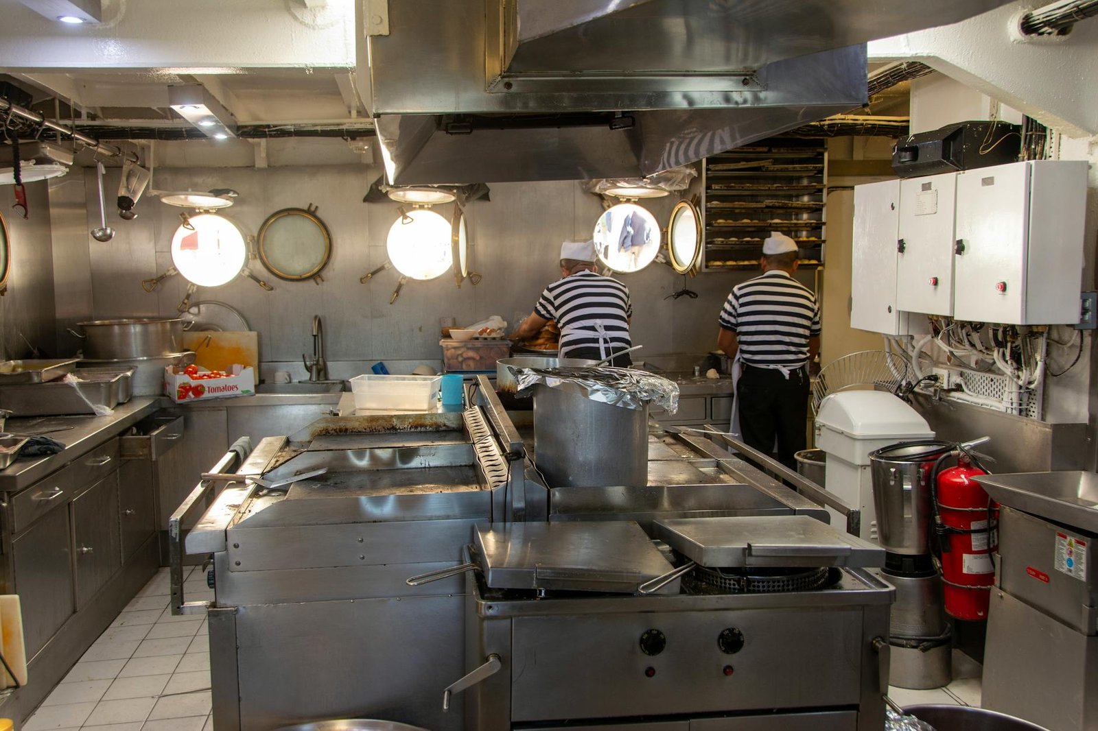 Two chefs in a ship's kitchen preparing meals with industrial equipment.