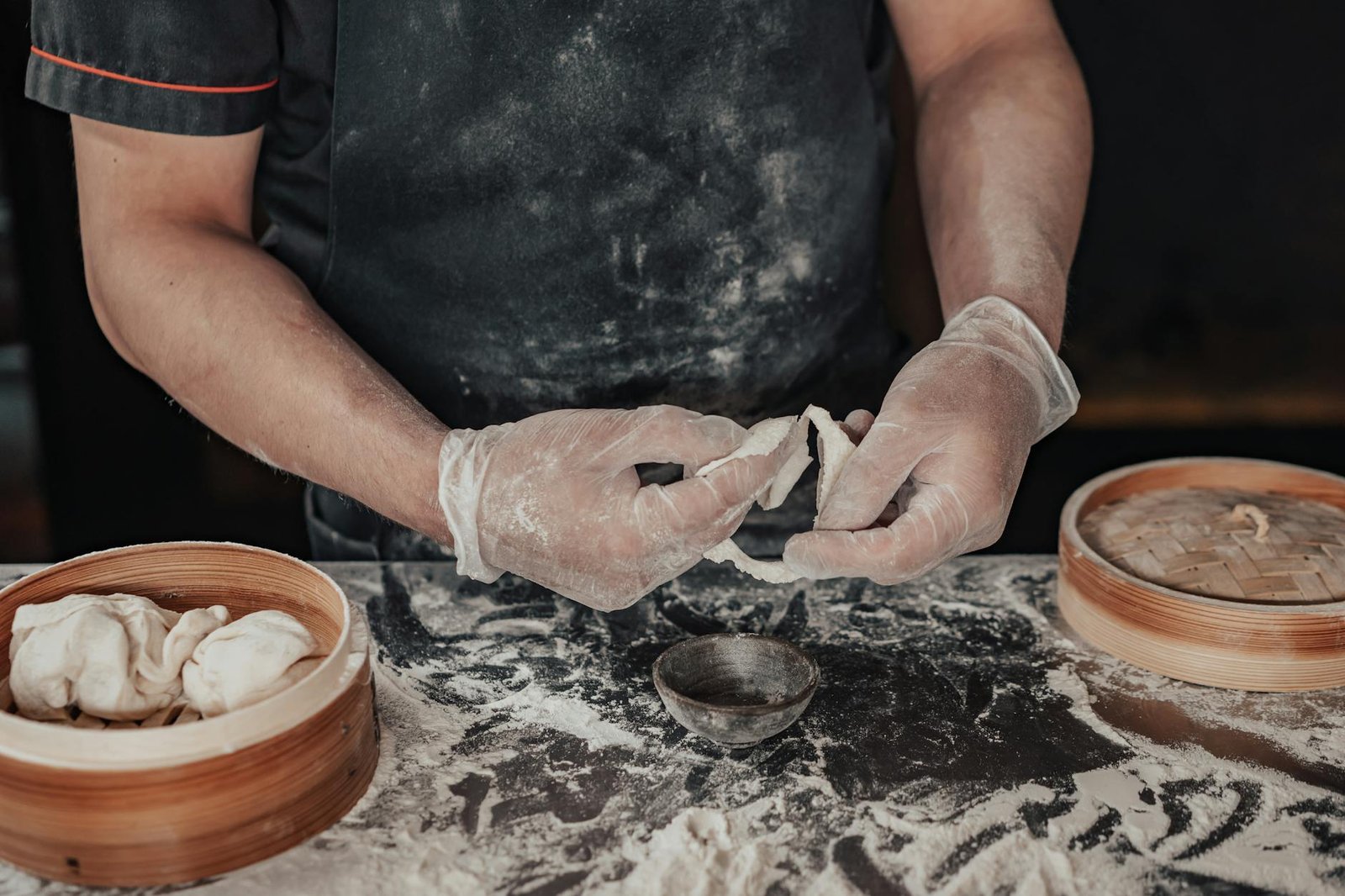 A chef wearing gloves expertly shapes dumplings made from dough using a bamboo steamer.