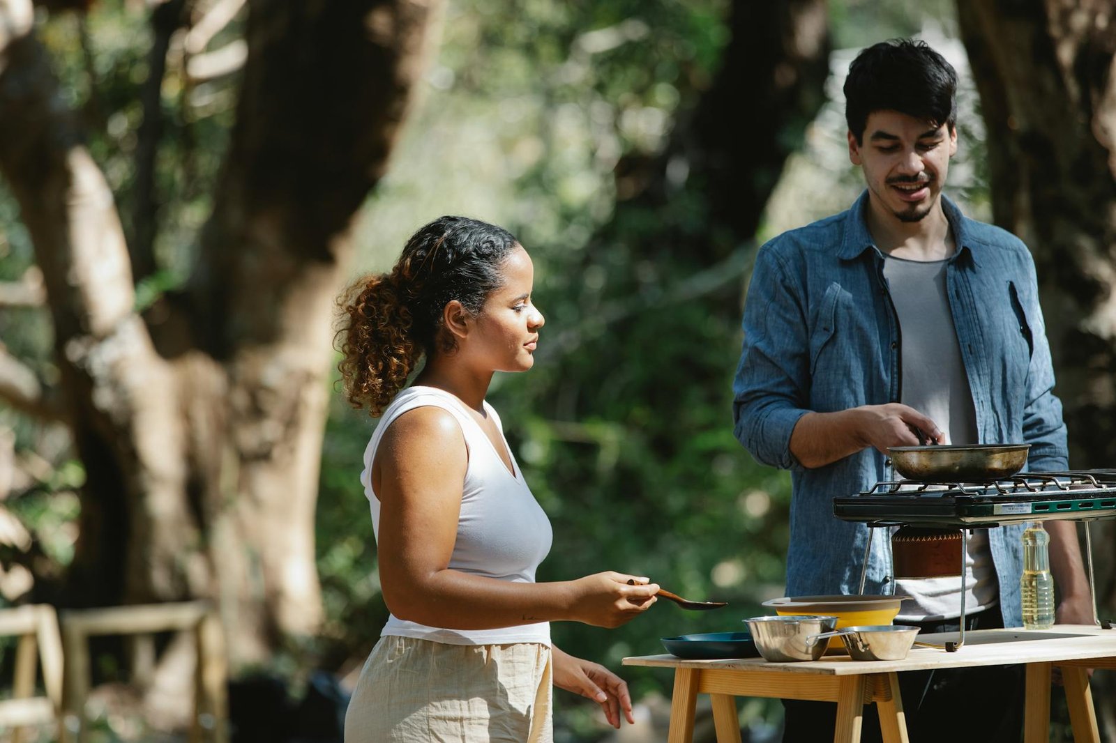 Focused multiethnic couple cooking on metal stove using skillet and spatula in woods