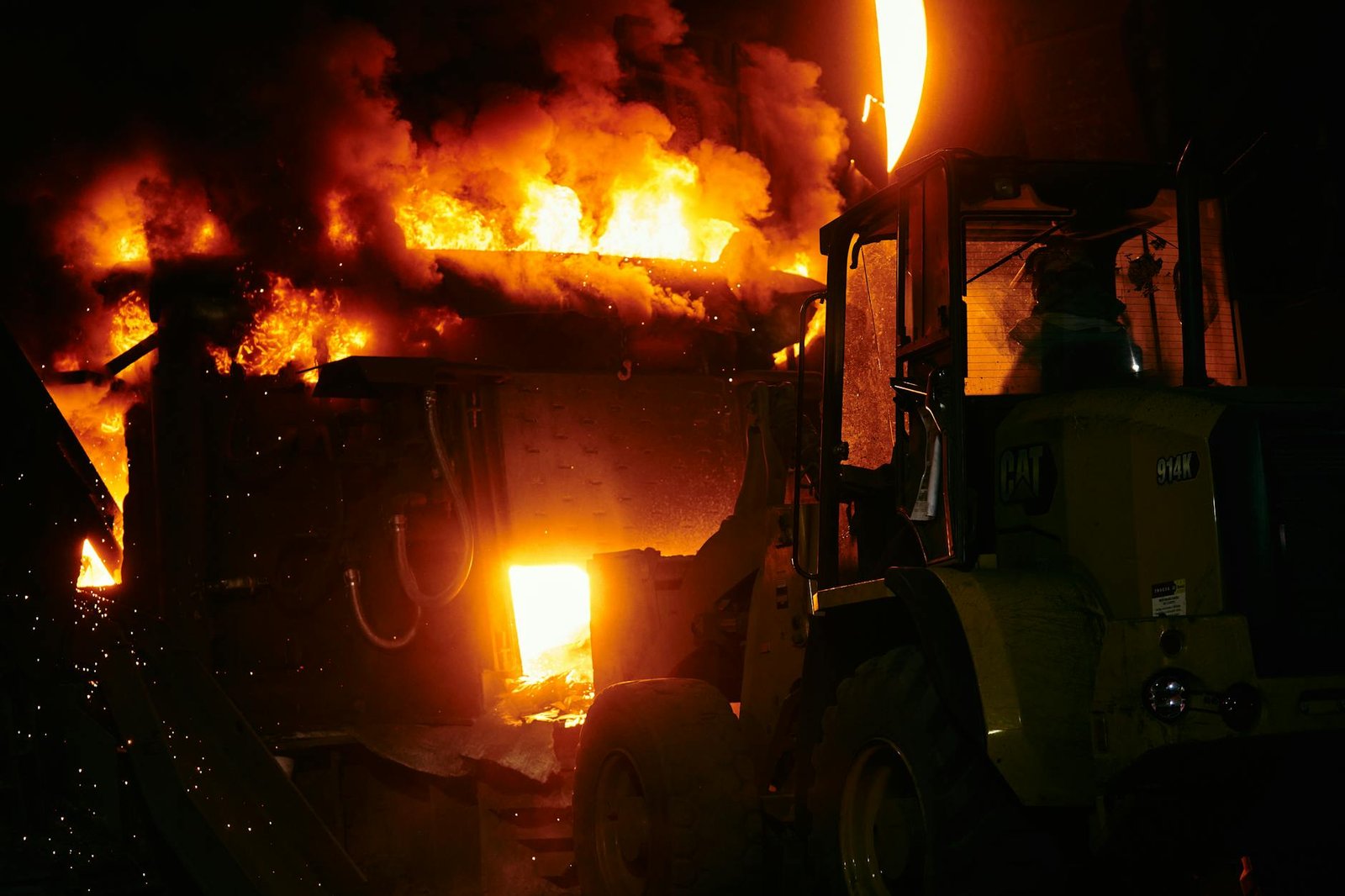 A powerful image of an intense industrial fire with a vehicle nearby, taken at night.