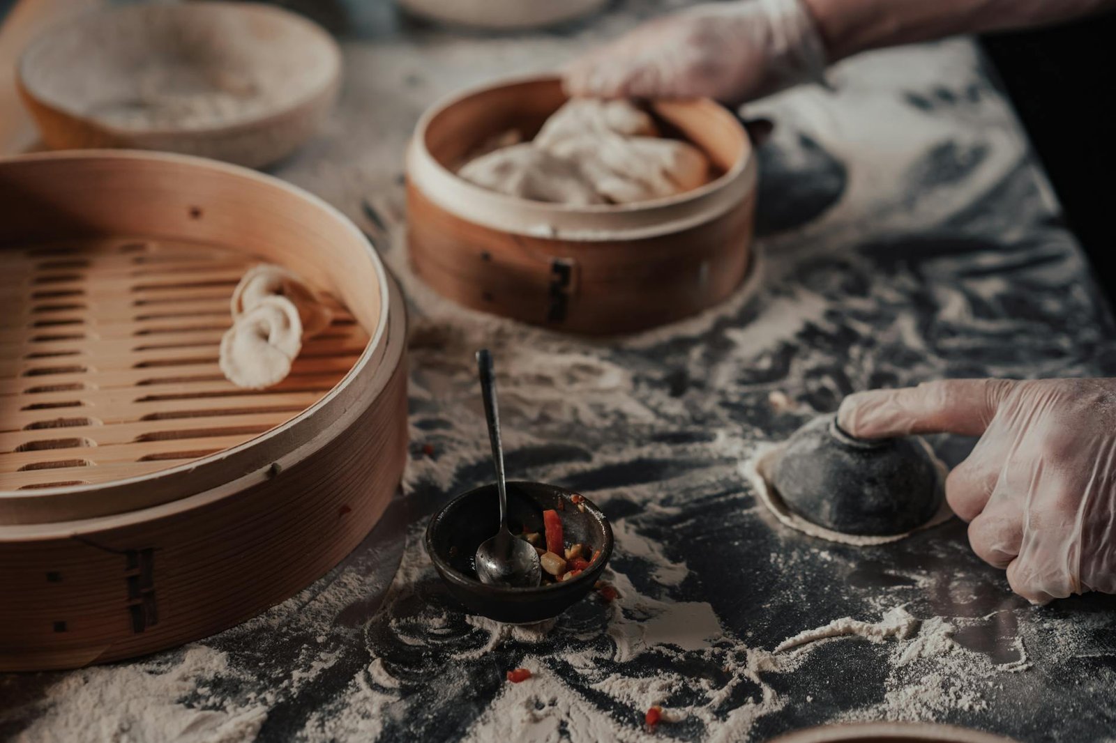 Close-up of hands making dumplings with bamboo steamers and flour in a traditional setting.